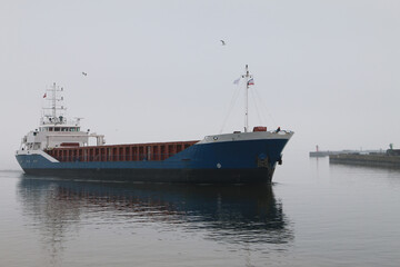 Fototapeta premium Cargo ship with open hold sails near harbor under calm skies. Seagulls fly overhead; breakwaters and lighthouses visible in the background. Peaceful maritime scene.