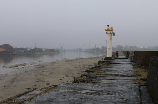 Foggy waterfront with concrete pier, white lighthouse, and person in orange vest walking toward it. Sandy shore, birds, and distant industrial cranes visible in misty background.