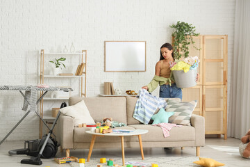 Tired mother with basket and scattered toys picking up dirty clothes for laundry in messy living...