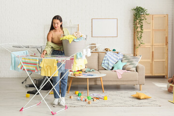 Happy mother with basket of laundry hanging clothes on dryer in messy living room
