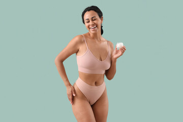 Young African-American woman in underwear with stretch marks and jar of cream on green background