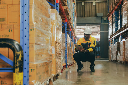 Black male salesperson wearing hard hat sits inspecting shelves using clipboard to record information about dietary supplement products in a warehouse waiting moved by a forklift.
