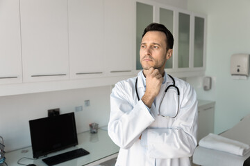 Serious male doctor standing in examining room deep in thought