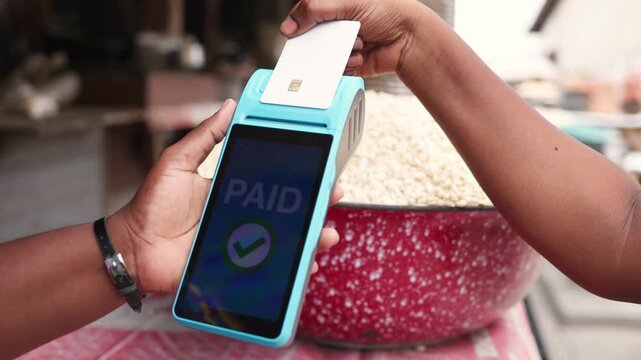 Contactless payment success, customer makes a transaction with a credit card via blue point of sale, POS terminal in a bustling outdoor African local traditional grains market.