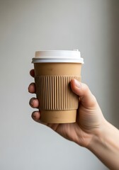 A light beige disposable coffee cup with a white lid is held in a hand against a plain gray background.