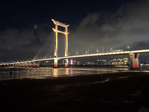 Foto noturna em Guangzhou, Guangdong (China) mostrando ponte estaiada iluminada sobre o Rio das P&eacute;rolas (Zhujiang), com skyline urbano ao fundo e reflexos na &aacute;gua.