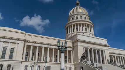 Obraz premium A beautiful white building with columns, a gilded dome, and a spire against a blue sky, clouds. Sculptures at the entrance. Old street lights. The inscription Capitol on the facade. Havana. Cuba