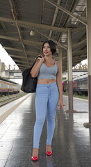 Fototapeta premium Asian woman in red heels and light blue jeans stands on a train station platform carrying a large black travel tote