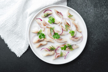Marinated herring slices with red onion and herbs on plate with napkin on dark background