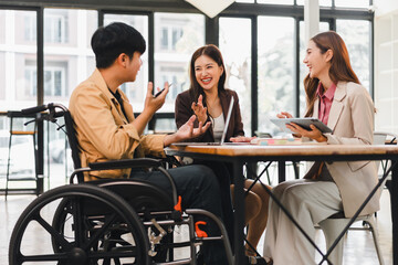 Young man in wheelchair disability meeting with two female colleagues around table discussing laptop and notes