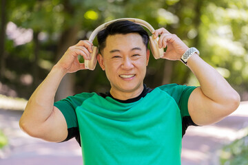 Happy young Japanese man put on wireless modern headphones earphones. Runner jogger guy sport male run running in park outdoors enjoy music listening song audio jogging healthy lifestyle fitness.