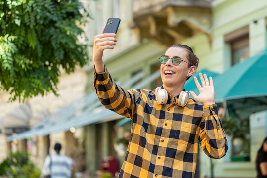 Cheerful man blogger taking selfie on smartphone, communicating video call online with subscribers or family friends, recording stories for social media vlog, waving hello. Guy on urban city street. - Powered by Adobe