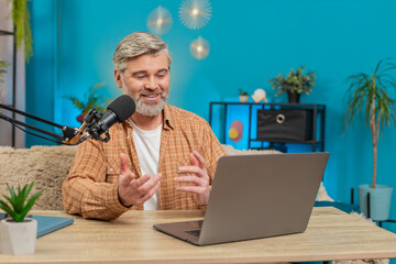 Caucasian mature man blogger speaking into studio microphone interacting with viewers subscribers during live online broadcast stream vlog. Middle-aged guy sitting at home table gestures, feedback