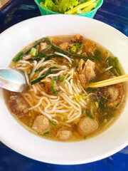 A bowl of noodle soup with a clear broth, containing noodles, pieces of meat, fish balls, green onions, and other garnishes. A spoon and chopsticks are placed in the bowl, and a basket of fresh greens