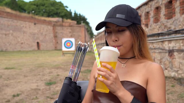 Beautiful young Asian woman in a cap enjoys a cold drink while standing at an archery range, holding her quiver filled with arrows