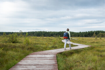 Rear view of woman traveler with basket of mushrooms, returning from forest along wooden path through wooded, elevated peat bog in autumn. Active lifestyle on nature