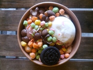 Close-up of a bowl of vanilla ice cream served with colorful cereal balls, chocolate cookies, and rainbow sprinkles. Fun and vibrant sweet dessert presentation on a wooden background.