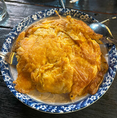A Thai-style omelet, likely Kai Jeow. The omelet is golden brown, fluffy, and appears to have a slightly crispy texture. It's served on a white plate with a blue floral pattern, with a fork.