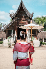 A woman in traditional Thai clothing holds a parasol, standing before an ancient temple under a bright blue sky.