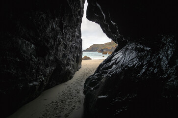 Narrow cave like passage between large dark rocks on smooth sand beach at Kynance Cove, footprints over ground, ocean in background