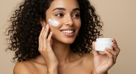 Woman applies face cream smiling holding jar against a neutral background.