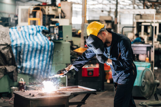 worker is actively engaged in factory.wearing a dark blue jumpsuit and a yellow hard hat, with a welding helmet covering his face for protection. Sparks creating a bright flash of light with metal