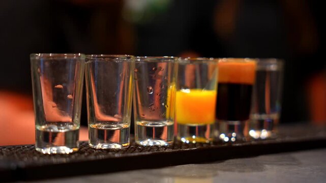 Bartender pouring colorful shots into row of glasses on bar counter in dim light creating nightlife and party atmosphere