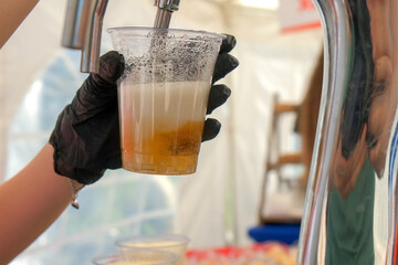 Bartender pouring fresh golden alcohol beer from tap in plastic cup in street cafe. Showcasing the process of serving a refreshing beverage at a bar or outdoor event. Foam rises as drink fills cup.