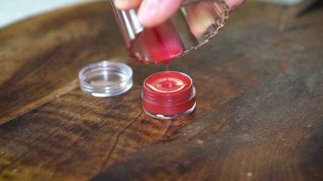 Making homemade natural lip balm with red pigment in transparent jar on dark wooden background. DIY beauty skincare concept with organic ingredients, eco-friendly cosmetics, and handcrafted style.