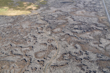 aerial view of a vast volcanic lava field, with a winding road cutting through the rugged, textured terrain. The high-angle perspective highlights the desolate beauty and unique patterns of the landsc