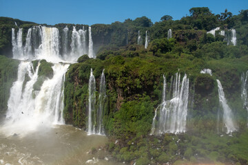 Fototapeta premium Iguazu Falls, showcasing its multiple, powerful cascades and a vibrant rainbow arcing through the mist. The shot captures the vast scale and stunning natural beauty of the site, making it ideal for to