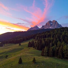Stunning mountain landscape at sunset. Lush green hills meet a dark forest at the base of a majestic mountain range, bathed in vibrant sunset colors