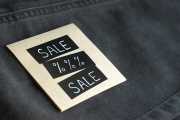 SALE note in denim jeans closeup. Square paper label with SALE text placed over black denim fabric, closeup minimal background for retail.