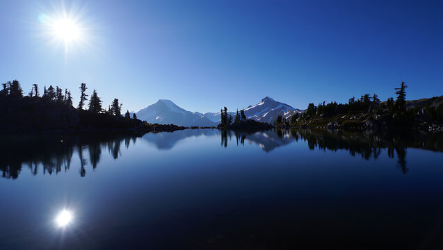A serene mountain lake reflects snowcapped peaks and evergreen trees under a bright, sunlit blue sky