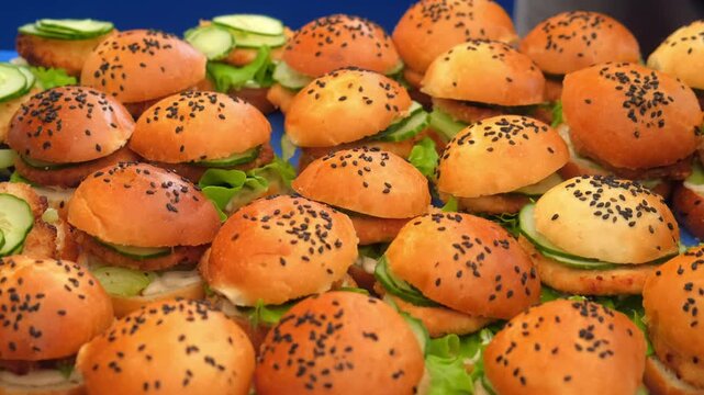 Displaying cooking rows of delicious mini burgers getting ready for catered event. Delicious burgers with sesame seed buns, fresh lettuce, cucumber slices and meat. Catering, street fast food snack.