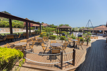 Bright, sunny view of an inviting empty outdoor patio at a cafe or restaurant, with wooden tables and chairs set up under a pergola, surrounded by lush green plants and landscaping