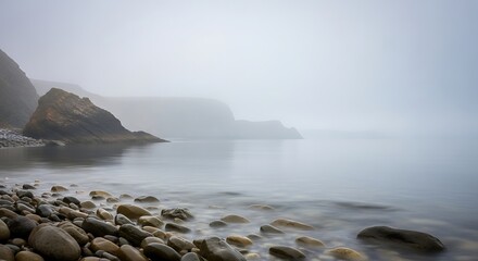 A tranquil coastal scene, shrouded in a soft, light gray mist, showcases smooth stones along the water's edge, with muted hills and rocks visible in the distance.
