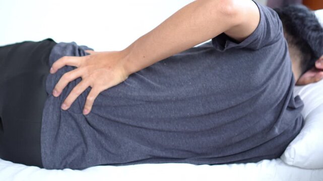 Exhausted Asian man lying in bed with severe lower back pain caused by poor sleeping posture, holding his back in discomfort. He suffers from muscle strain, stiffness, and body aches after waking up
