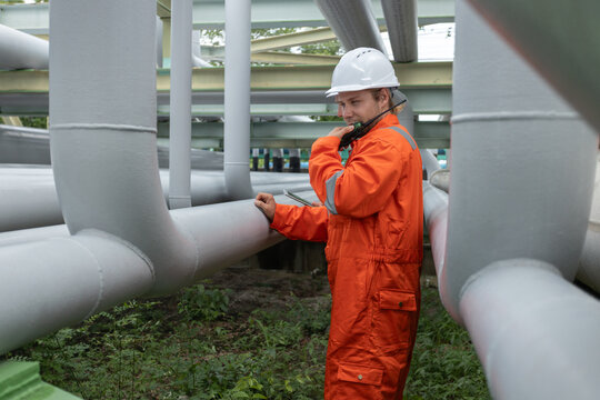 Industrial engineer in orange safety uniform and hard hat inspects pipeline system in field. Communication via walkie-talkie and digital tablet ensures safety and maintenance. - Powered by Adobe