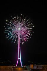 A striking modern light sculpture, resembling a dandelion or fireworks, illuminates the night sky. Its colorful stand and bright points of light create a vibrant display overlooking Lake Como.