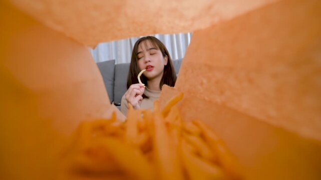 Creative perspective shot of Asian woman reaching into a bag of french fries, symbolizing junk food indulgence, casual lifestyle, and unhealthy eating habits.