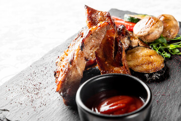 Grilled meat ribs with roasted vegetables, baby potatoes, and red sauce on a dark slate board. Rustic restaurant-style food presentation, top view, close-up