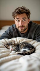 Obraz premium Young man with beard, wavy brown hair relaxing on bed, kitten curled on sweater, pets nearby. Concept of cozy moment, kitten curled on sweater in warm atmosphere.
