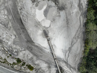 Industrial quarry crusher facility from above with conveyor belts and raw material piles, surrounded by erosion patterns and forest edge, symbolizing resource extraction and environmental pressure.