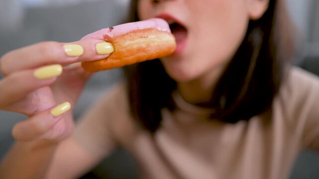 Young woman eating a donut indoors, close-up view showing hand and face while enjoying sweet food, representing casual lifestyle and junk food indulgence.
