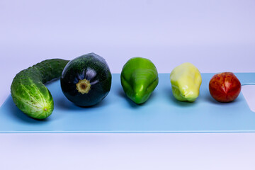 A clean studio shot of a colorful assortment of vegetables—a cucumber, zucchini, two bell peppers, and a tomato—lined up on a bright blue cutting board.

