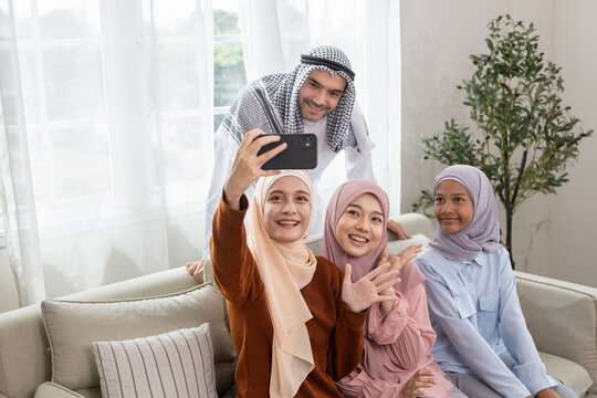 Happy Muslim family taking selfie with smartphone at home. Parents and children smiling together on sofa, concept of Islamic lifestyle, bonding, modern technology and cultural tradition.
