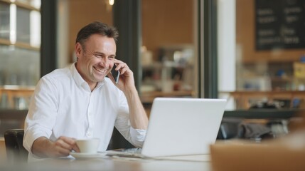 Fototapeta premium Man in white shirt is smiling while talking on phone in open office workspace, holding coffee cup and using laptop. atmosphere is professional and relaxed