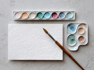 Artist's watercolor set with a paintbrush and blank textured paper on a grey concrete surface.