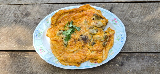 Thai style omelette served on the wooden table : Omelette with fresh oysters fried in oil until crispy, sprinkled with pepper and coriander. Fried beaten egg with oysters, crispy flour, Eat with sauce